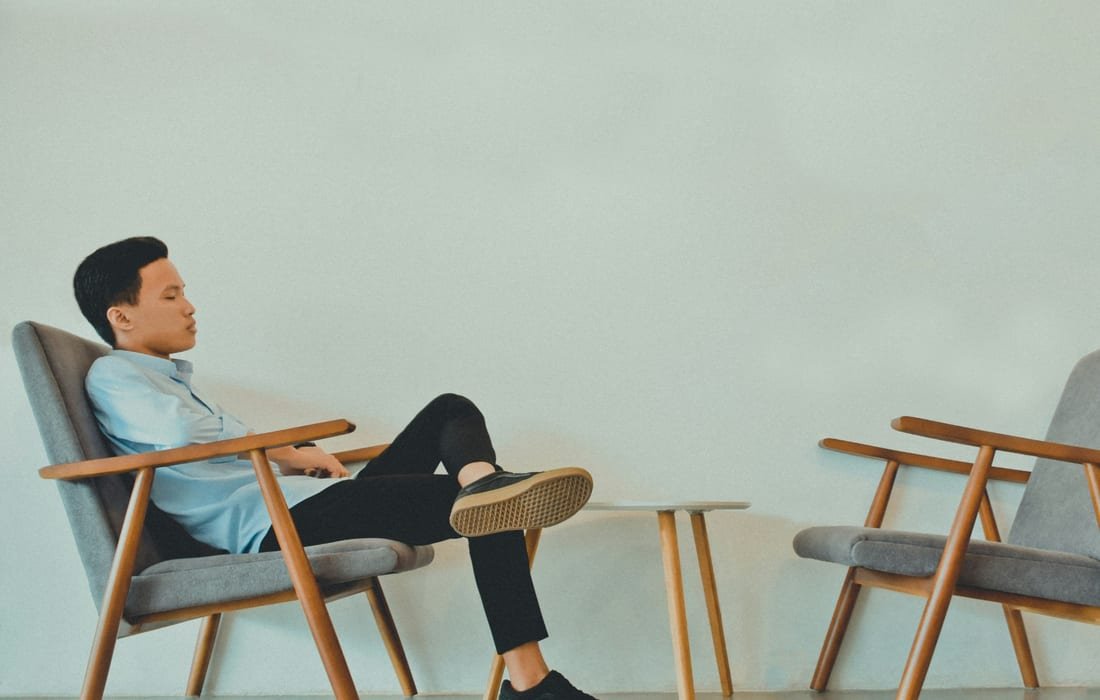 A man sitting back in a grey armchair with wooden arms, a small table and an empty second armchair beside him against a pale wall