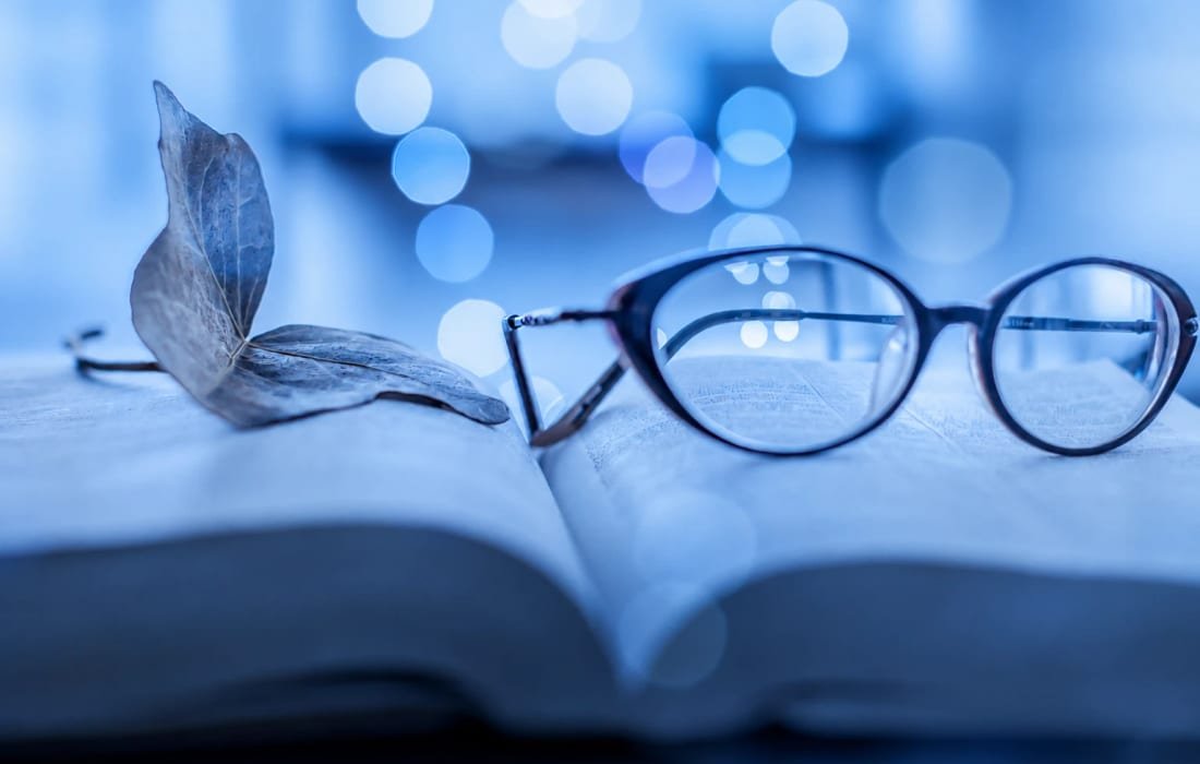 A pair of reading glasses and a dried leaf resting on an open book, in cool blue light with soft lights behind