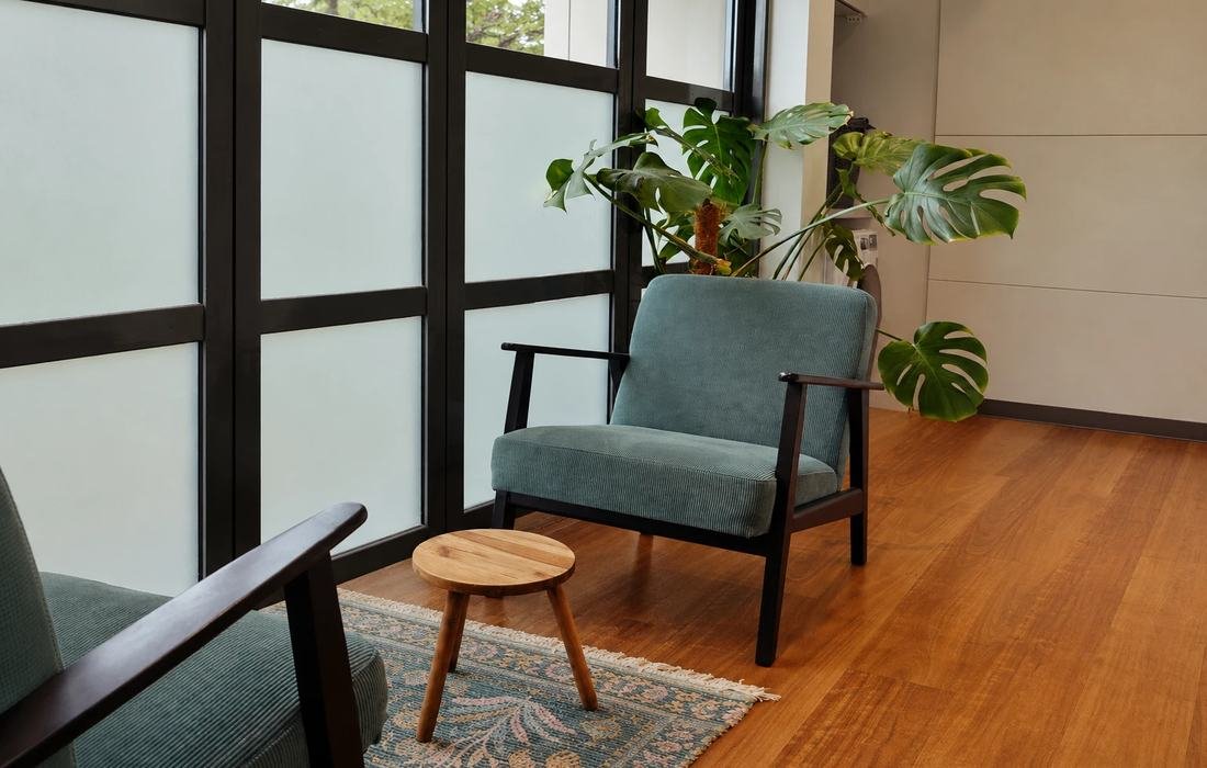 A green corduroy armchair beside tall frosted windows, a small round wooden stool on a patterned rug, and a monstera plant in the corner