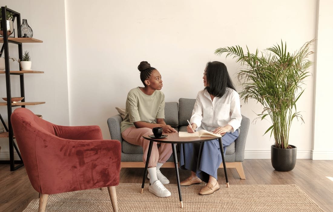 Two people talking on a sofa across a small round table, one writing in a notebook, a red armchair and a tall plant either side