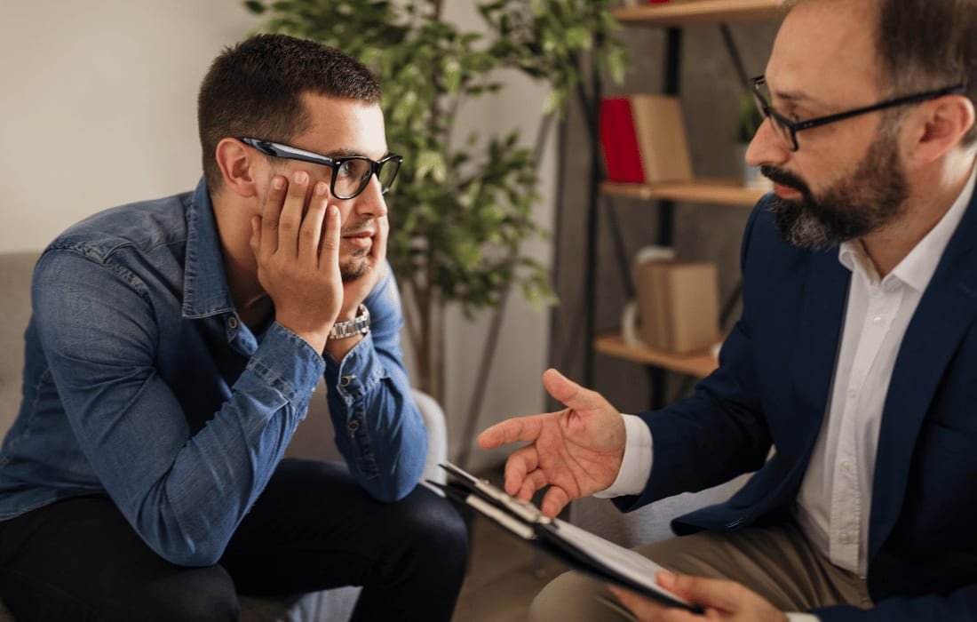 Two men in a room with bookshelves, one listening with a tablet on his knee, the other mid-sentence